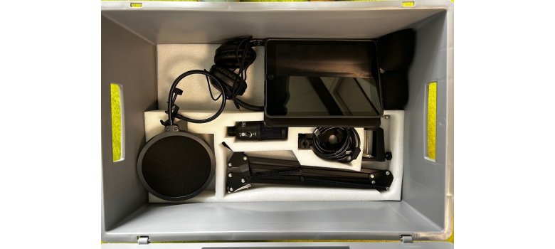 Eine Kiste ist zu sehen, in der die Untensilien für einen Podcast sind: Kopfhörer, Stativ, Mikrofon, Tablet.