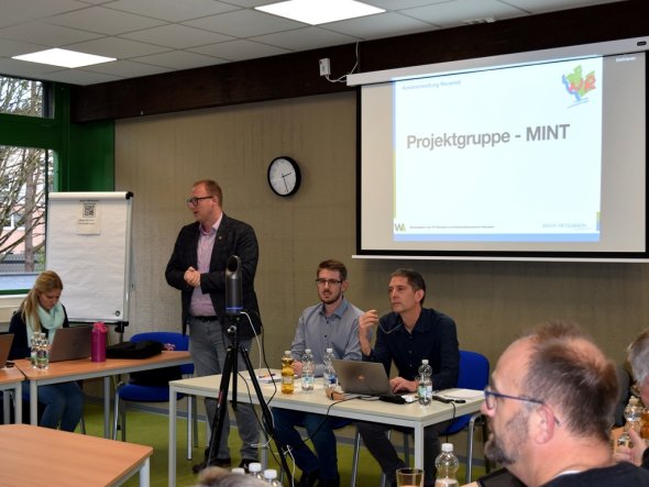 Zwei Männer sitzen hinter einem Tisch. Neben dem Tisch steht ein weiterer Mann und hält einen Vortrag. Im Hintergrund sieht man eine Leinwand auf der "Projektgruppe MINT" zu lesen ist.