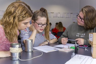 Drei Schülerinnen sitzen an einem Tisch und versuchen eine Aufgabe zu lösen. Auf dem Tisch sind 2 Blechdosen mit eine Kordel und eine Batterie mit einer kleinen Birne zu sehen.